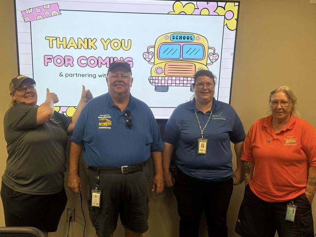 A group of four school transportation staff stand in front of a screen with a colorful school bus graphic and text that reads, “Thank you for coming & partnering with us.” The individuals are smiling, showing appreciation for their role in supporting students and families.