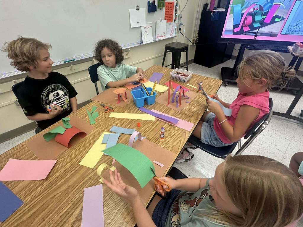  A high-angle view shows four children sitting at a long table in a classroom, engaged in an art project. They are cutting and gluing colorful paper strips, creating three-dimensional sculptures. A large monitor in the background displays an example of similar artwork.