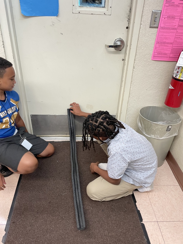 Two students sit on the floor by a door, setting up a foam tube ramp for a science experiment.