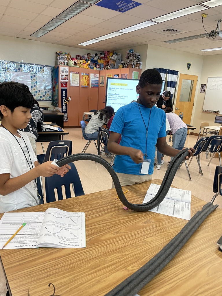 Two students at a classroom table hold a wavy foam ramp with lab papers in front of them.
