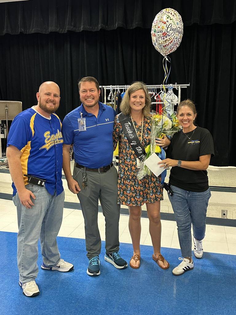  A full-body photo of four people standing on a stage with a dark blue curtain backdrop. From left to right: a bald man in a yellow and blue jersey, a man in a blue polo shirt, a woman in a floral dress wearing a "Teacher of the Year" sash and holding a bouquet, and a woman in a black t-shirt. They are all smiling.