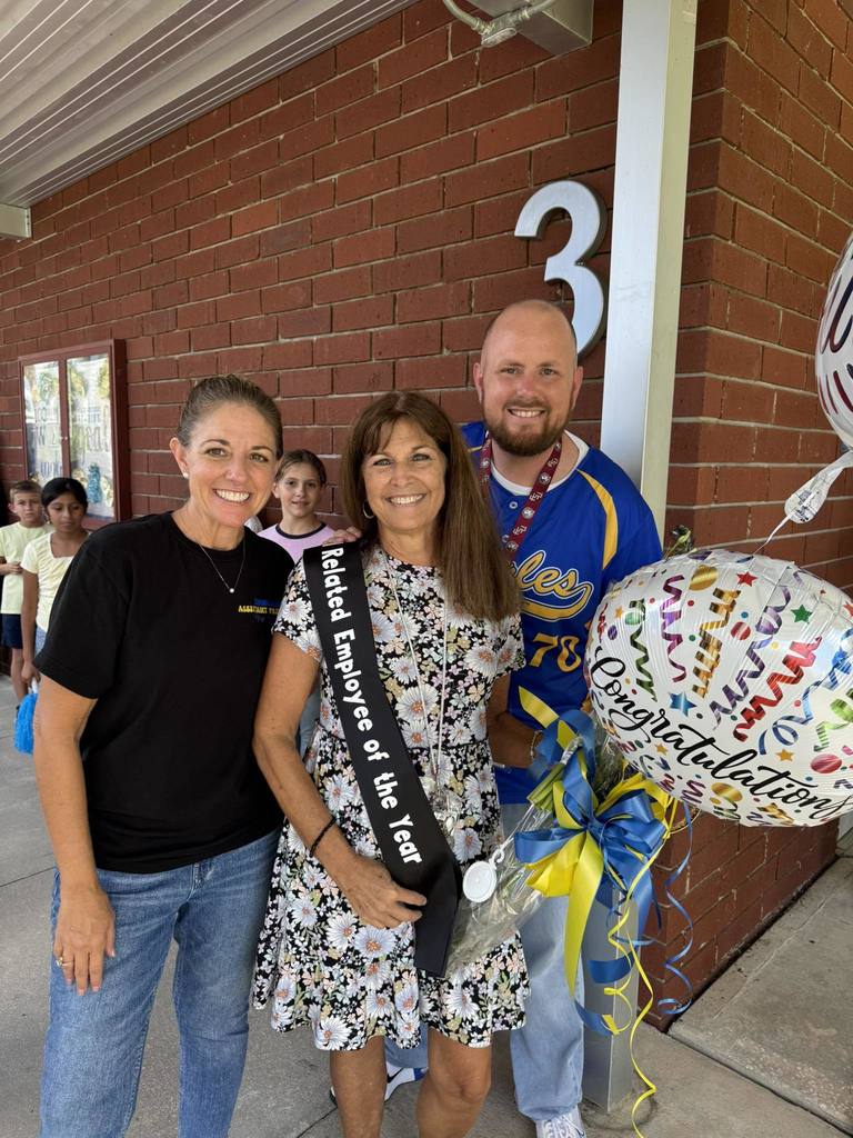  A woman in a floral dress and a "Support-Related Employee of the Year" sash stands between a woman in a black t-shirt and a man in a blue and yellow jersey. They are all smiling. The woman in the middle is holding a bouquet and a "Congratulations" balloon. In the background, two children are partially visible, and a brick wall with a large number "3" is seen.