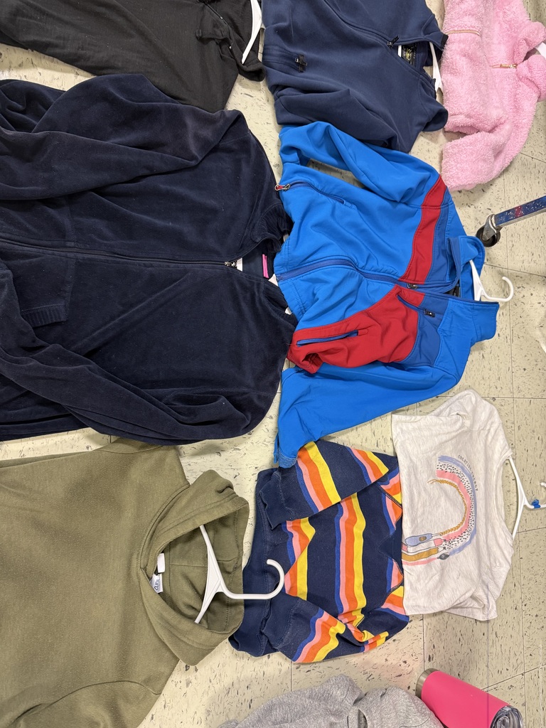 A variety of lost-and-found items, including jackets, water bottles, and lunchboxes, are laid out on a beige tile floor. The items are spread out in groups, with many colorful jackets and several water bottles with popular cartoon characters.