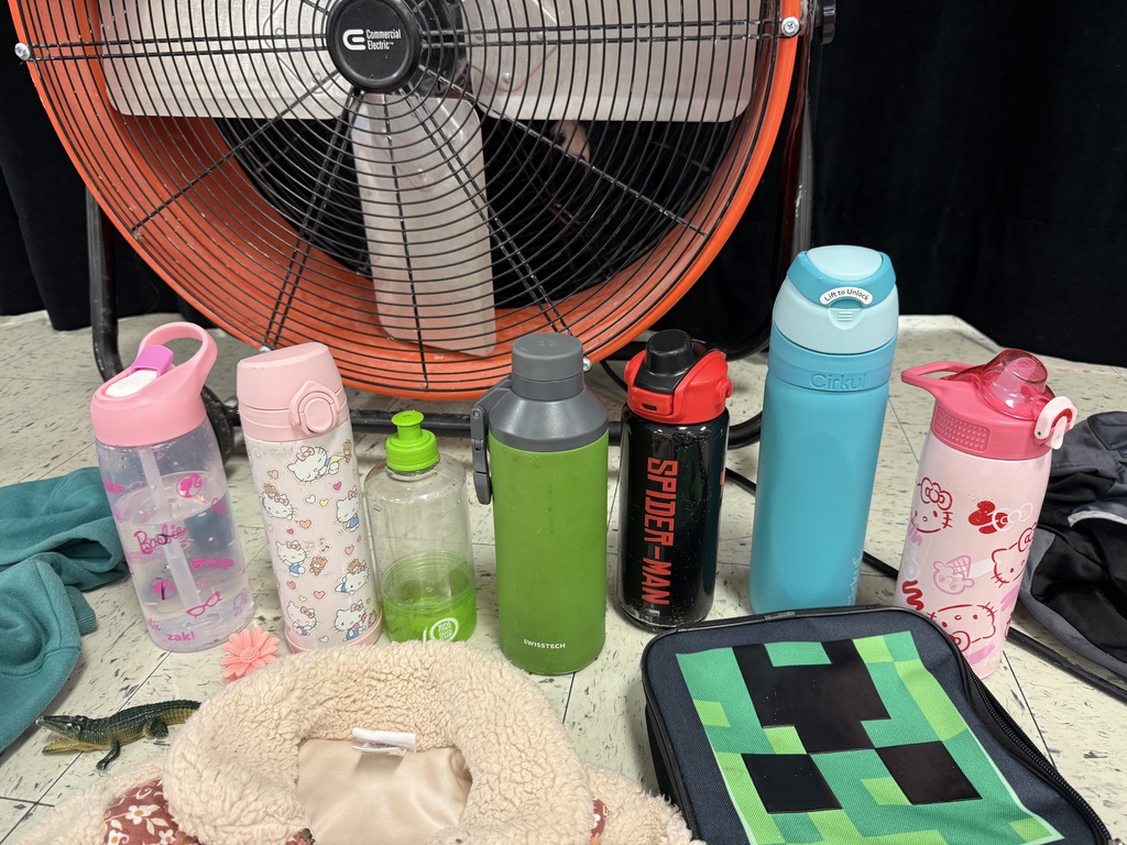  A variety of lost-and-found items, including jackets, water bottles, and lunchboxes, are laid out on a beige tile floor. The items are spread out in groups, with many colorful jackets and several water bottles with popular cartoon characters.