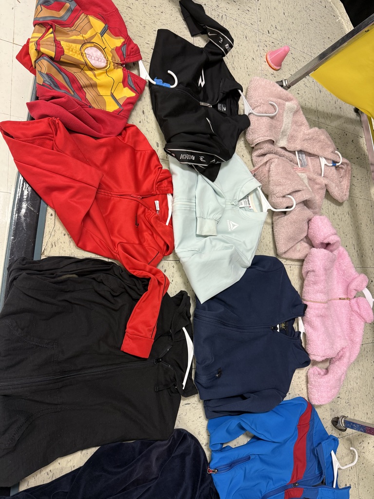  A variety of lost-and-found items, including jackets, water bottles, and lunchboxes, are laid out on a beige tile floor. The items are spread out in groups, with many colorful jackets and several water bottles with popular cartoon characters.