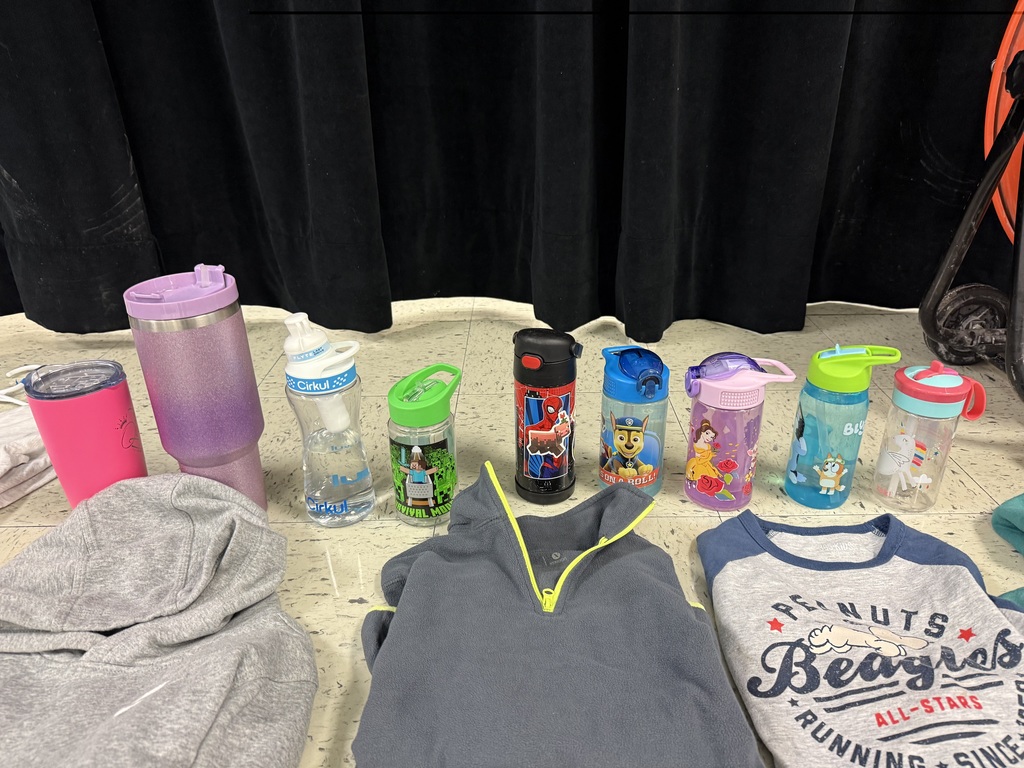  A variety of lost-and-found items, including jackets, water bottles, and lunchboxes, are laid out on a beige tile floor. The items are spread out in groups, with many colorful jackets and several water bottles with popular cartoon characters.