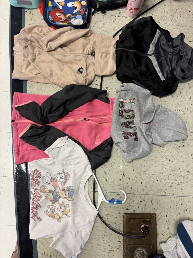  A variety of lost-and-found items, including jackets, water bottles, and lunchboxes, are laid out on a beige tile floor. The items are spread out in groups, with many colorful jackets and several water bottles with popular cartoon characters.