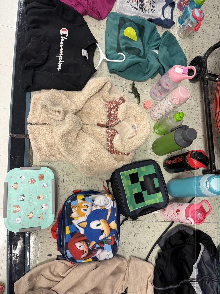  A variety of lost-and-found items, including jackets, water bottles, and lunchboxes, are laid out on a beige tile floor. The items are spread out in groups, with many colorful jackets and several water bottles with popular cartoon characters.