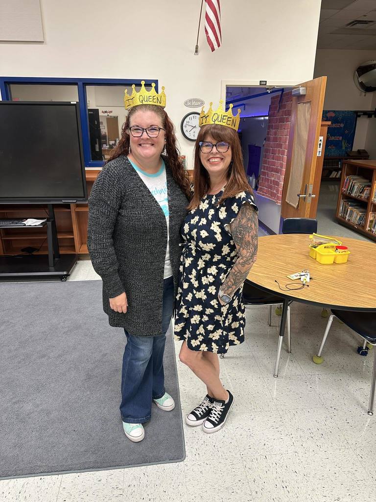  Two women stand side by side in a classroom. Both are smiling and wearing small gold crowns with "IT QUEEN" written on them. The woman on the left wears a gray cardigan, and the woman on the right wears a floral print dress.