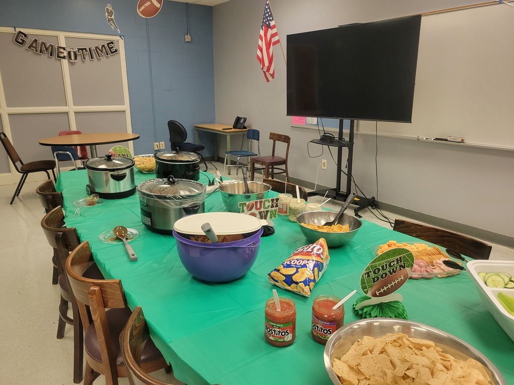 A long table is set with a green tablecloth and covered in food for a party, including two Crock-Pots, chips, dips, and a platter of food. A "GAMETIME" banner is visible in the background, along with a large TV screen and an American flag.