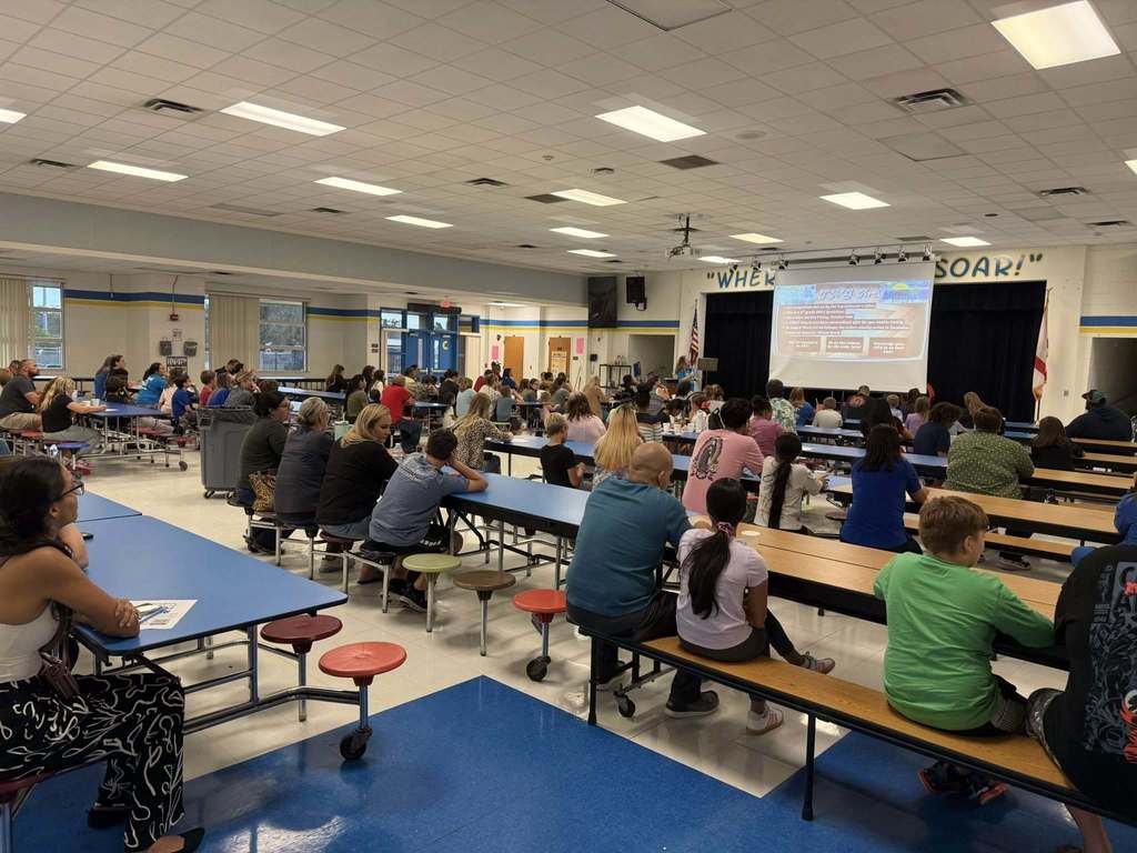  A large group of people, including adults and children, are seated at cafeteria tables with attached benches. They are facing a stage area where a presentation is being shown on a screen. The cafeteria has blue floors and white ceiling tiles.