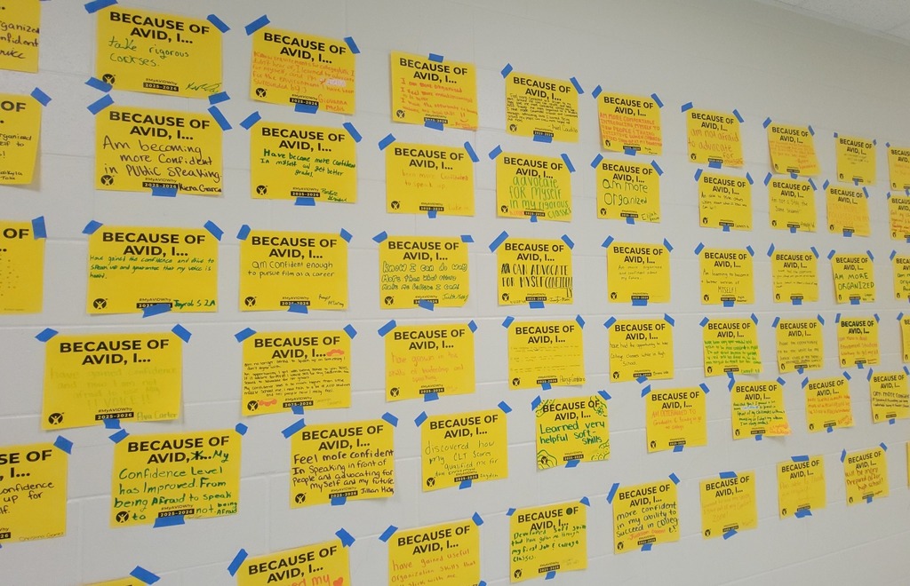 A white wall is covered in yellow sticky notes, each with a student's handwritten response to the prompt "Because of AVID, I...". The notes are attached to the wall with blue painter's tape, forming a large grid.