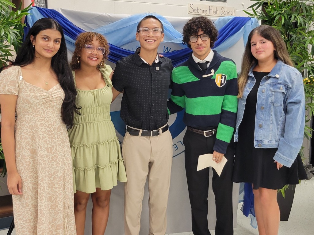 Five teenagers, three females and two males, are posing for a group photo in front of a white and blue banner with "Sebring High School" written on it. The students are dressed in semi-formal attire, with leafy green plants on either side of them.