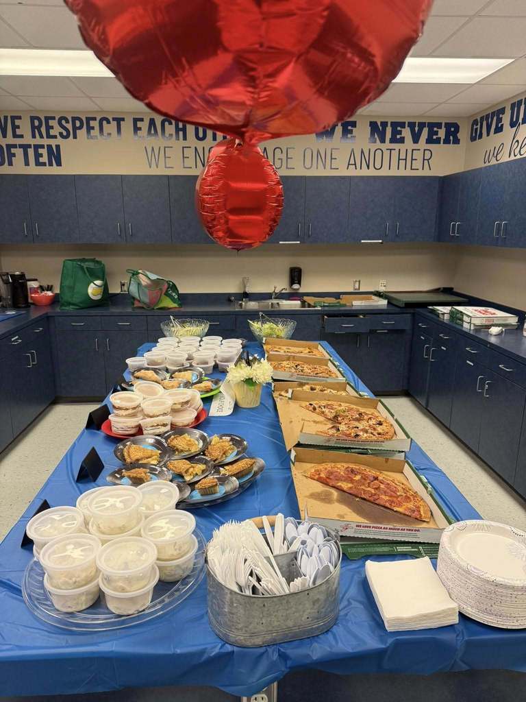  A blue-themed room, likely a staff lounge, is set up with a long table for a meal. The table is covered in a blue cloth and holds several pizza boxes, individual containers of food, silverware, and paper plates. Blue cabinets line the wall, above which is a motivational quote. A large red balloon hangs overhead.