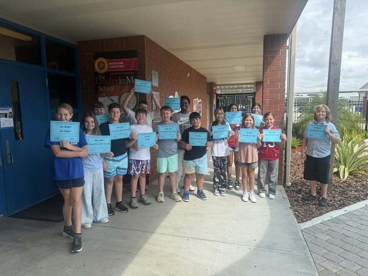  A group of about twelve elementary school students is standing outside a brick building under an awning, holding light blue certificates. They are smiling and posing for a photo. A sign on the building identifies the location as "Woodlands Elementary: The Leader in Me School."
