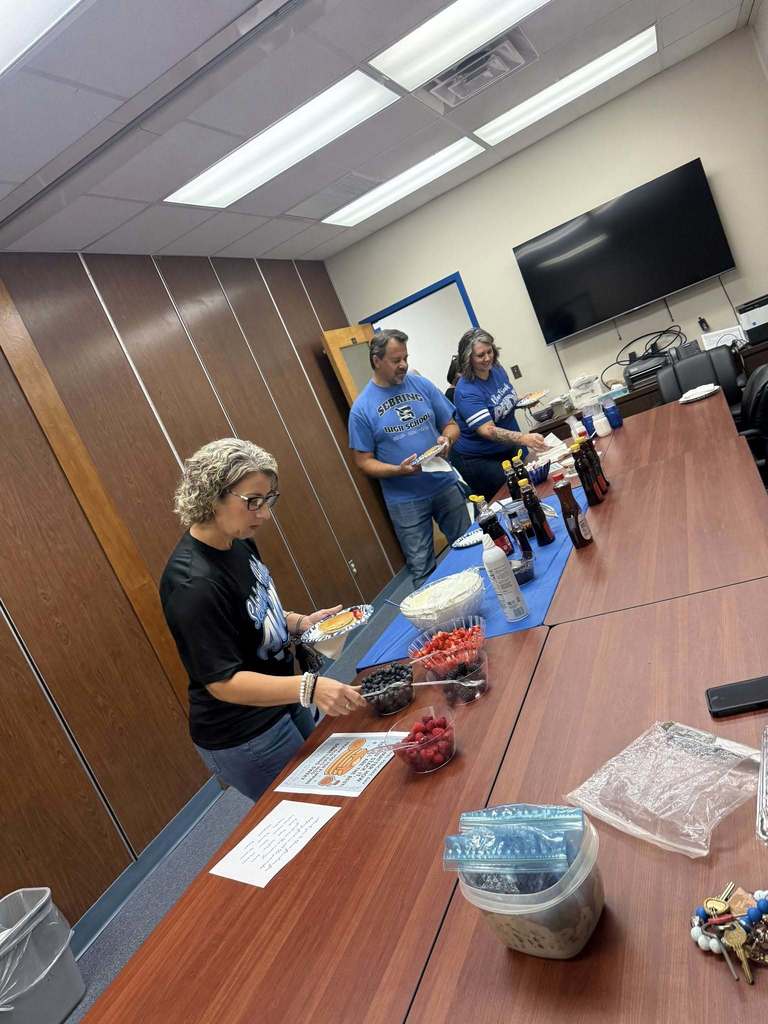  Three adults are standing behind a long wooden table setting up a breakfast spread in a conference room with wood-paneled walls. The table holds a variety of toppings, including bowls of fresh berries and whipped cream, syrup bottles, and plates of pancakes. Two of the adults are wearing blue Sebring High School shirts.