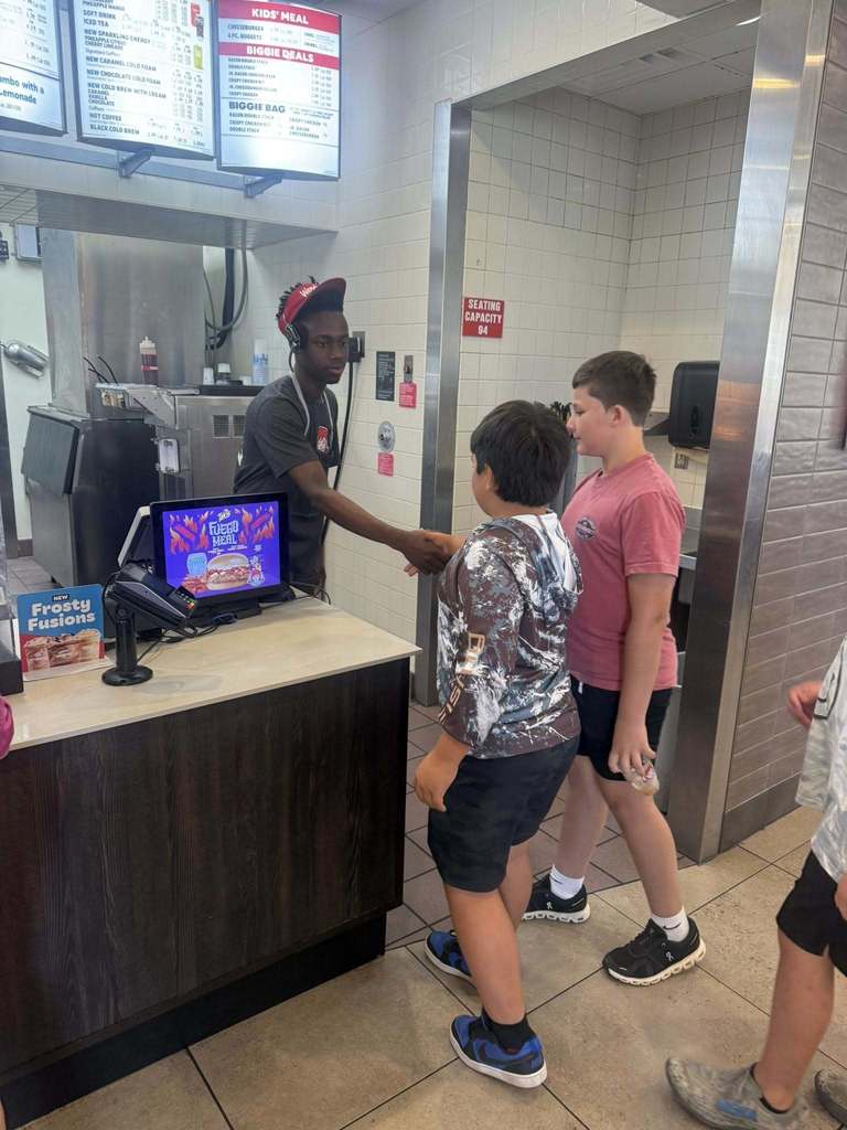  A food service worker wearing a headset and uniform is standing behind a counter, shaking hands with one of two young boys. A cash register screen is visible on the counter. The background includes menus and a sign indicating the seating capacity.