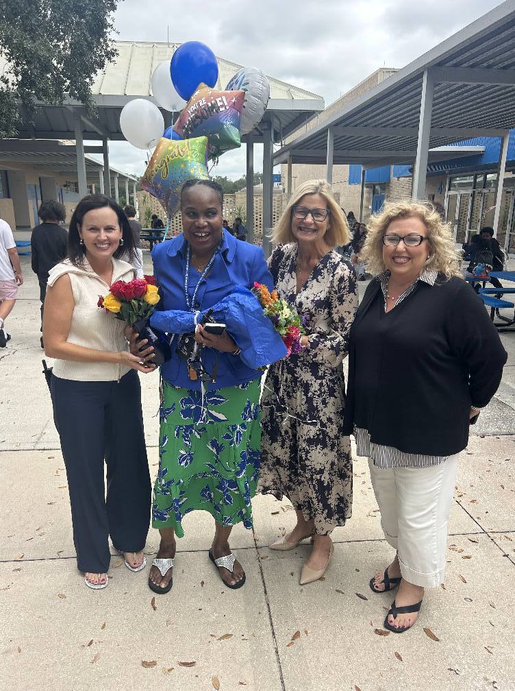  A group of four women are standing outdoors in front of a covered walkway. The woman second from the left is holding a bouquet of flowers and a cluster of celebratory balloons. All four women are smiling and posing for the camera.