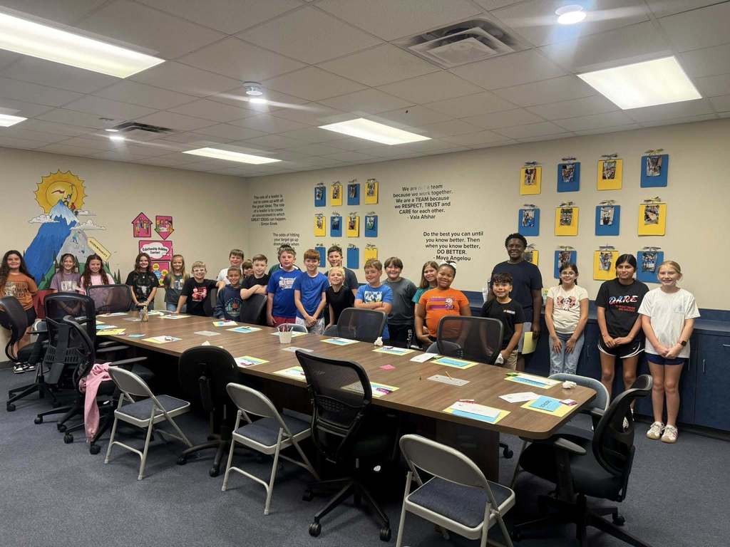  A long conference room table is surrounded by chairs. About 20 elementary school students are standing behind the table, smiling for a group photo. The walls are decorated with a mural and inspirational quotes, including posters with the text "We are a TEAM."