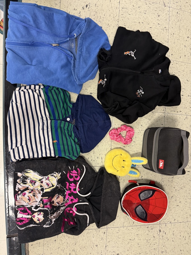A flat lay of multiple items, mostly clothing and accessories, on a tiled floor. Items include a blue zip-up hoodie, a black hoodie with small white graphic accents, a striped shirt (white/black and green/blue), a black lunch box, a Spiderman mask-shaped lunchbox, a pink scrunchie, and a yellow smiling bunny coin purse.