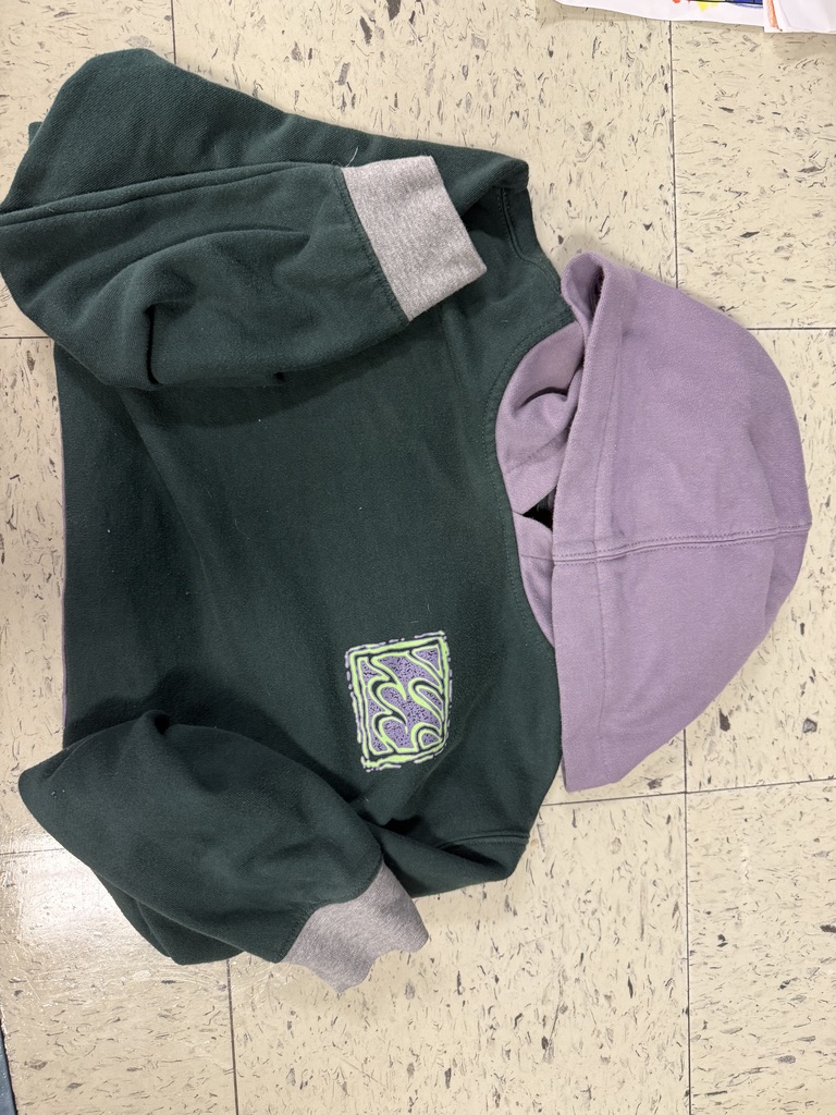 A dark green pullover hoodie with light gray cuffs and a large light purple hood. The chest features a small square patch with a green and purple swirling pattern.