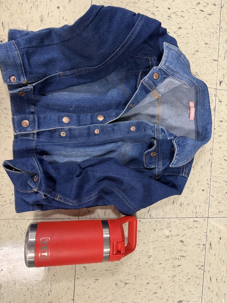 A blue denim jacket with copper-colored buttons laid out next to a large, red thermos with a handle, which appears to be a Yeti brand bottle.