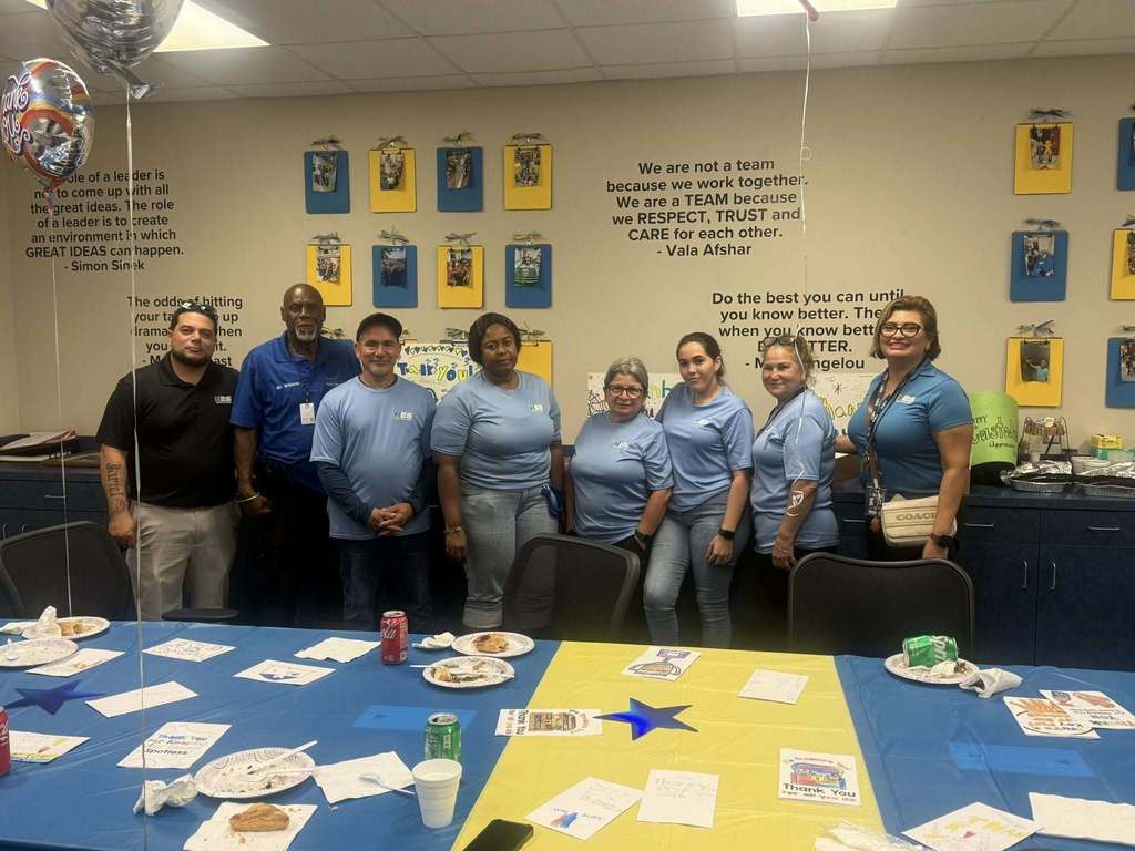  A diverse group of eight people, wearing light blue shirts, stand behind a table with snacks and drinks. Balloons and motivational quotes decorate the office wall behind them. They appear to be celebrating an event.