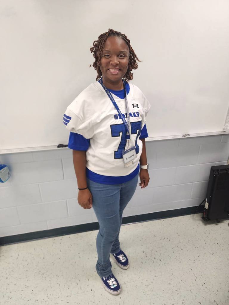  A smiling woman stands indoors in front of a whiteboard, wearing a white and blue football-style jersey with the number 77 and blue jeans.
