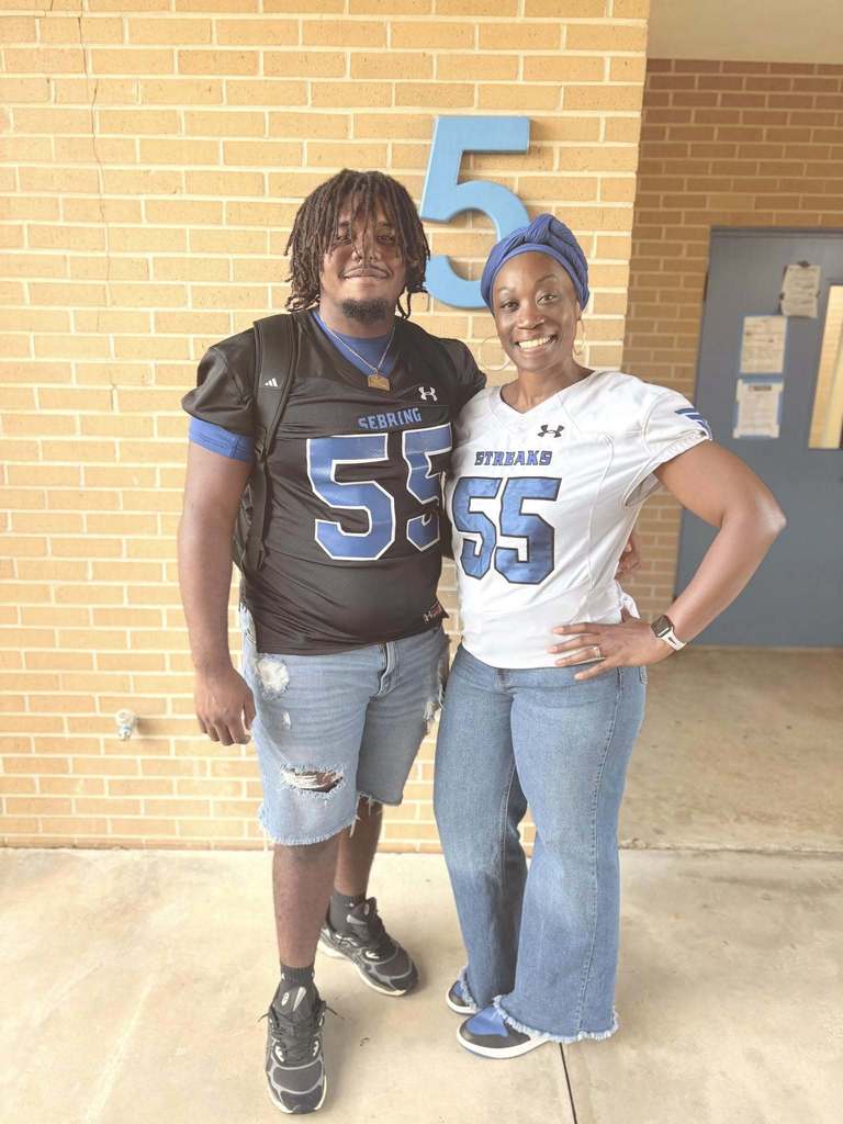 A young man wearing a black football jersey with number 55 and a woman in a white jersey with number 55 stand next to each other smiling outside. 