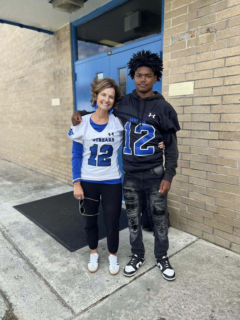A smiling woman and a young man stand outdoors in front of a brick building. They are both wearing football-style jerseys with the number 12. The woman wears a white jersey, and the man wears a black hooded jersey.