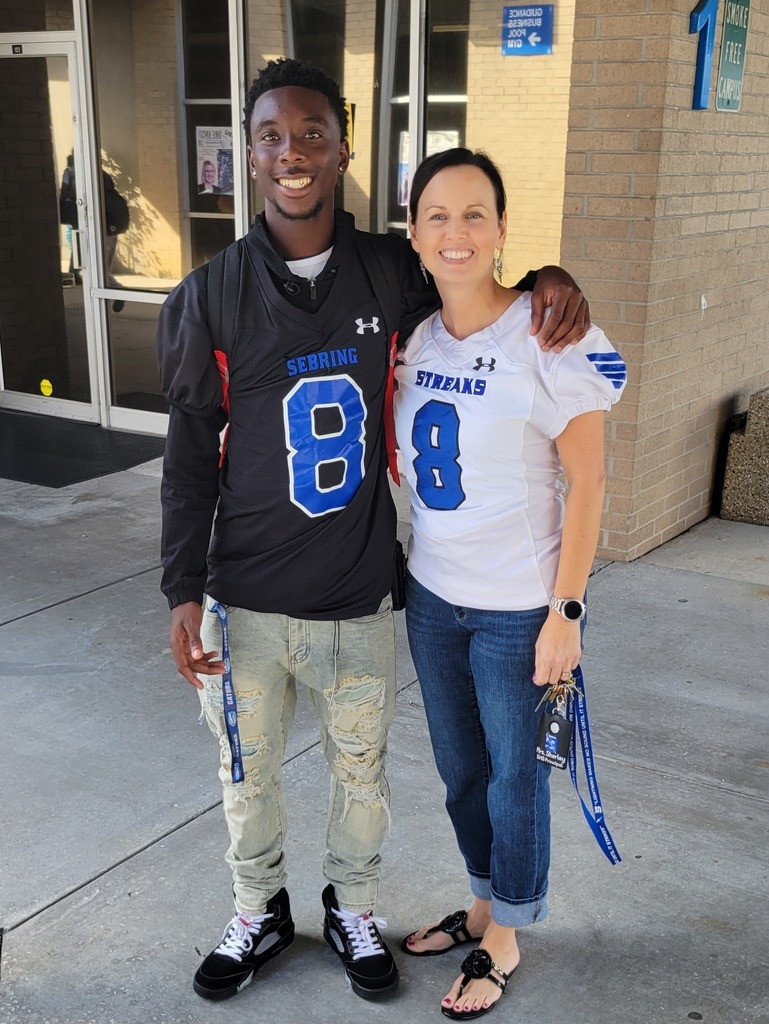  A young man in a black football jersey with number 8 and a woman in a white jersey with number 8 stand smiling, arms around each other's shoulders, outside a brick building.