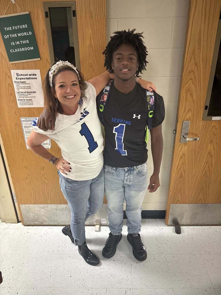 A woman in a white football jersey and a young man in a black jersey, both with the number 1, stand in a hallway. The woman has her arm over the man's shoulder.