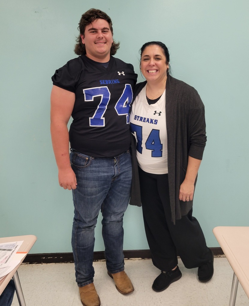 A young man wearing a black football jersey with number 74 and a woman in a white jersey with number 74 stand in a classroom with a light blue wall.