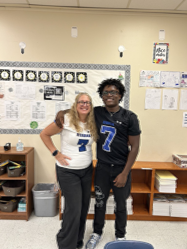 A woman in a white jersey and a young man in a black jersey, both featuring the number 7, stand in a classroom with colorful bulletin boards behind them.