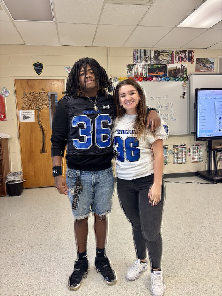 A young man in a black football jersey and a woman in a white jersey, both with the number 36, stand in a brightly lit classroom.