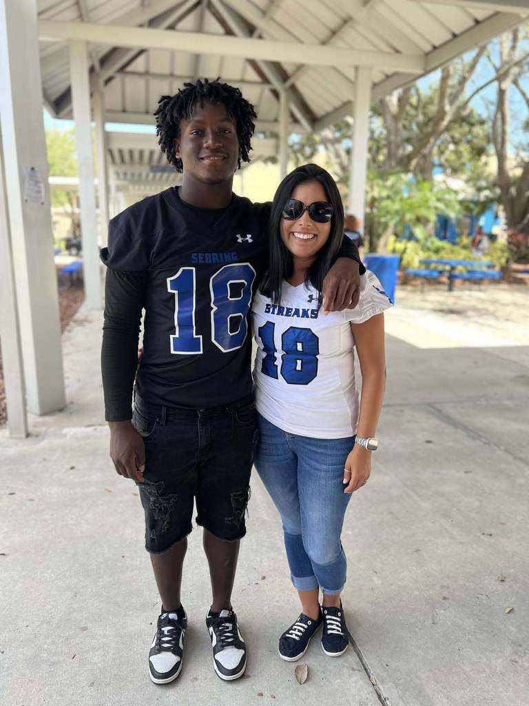 A young man and a woman, both wearing jerseys with the number 18, stand smiling under an outdoor awning. The man wears a black jersey and the woman wears a white one.