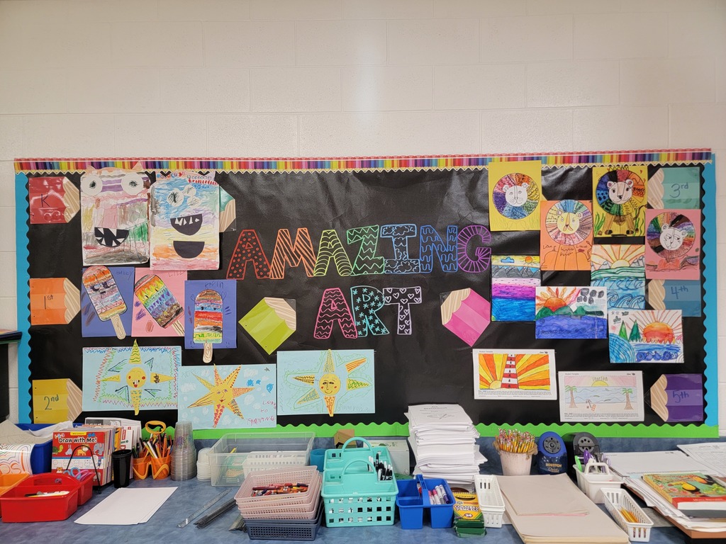A classroom art display board titled “Amazing Art” showcases colorful student artwork from Memorial Elementary. Pieces include drawings of monsters, popsicles, suns, lions, landscapes, and lighthouses, arranged by grade levels from 1st through 5th.