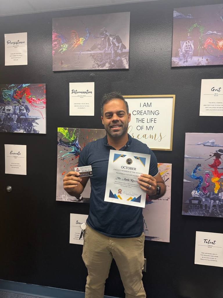 A smiling man in a navy shirt stands against a black wall covered with motivational art and signs. He holds a "Faculty Member of the Month" certificate for October and a small card.