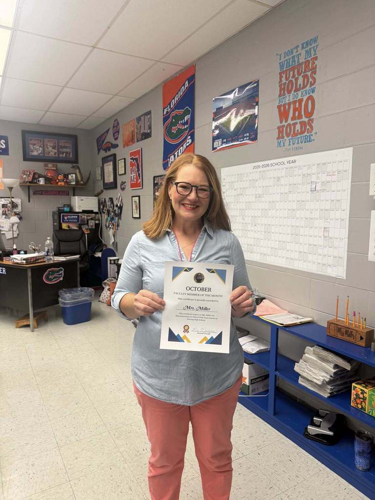  A smiling woman wearing a denim shirt and coral pants stands in an office holding a "Faculty Member of the Month" certificate for October. University of Florida Gators memorabilia and a calendar are on the wall behind her.