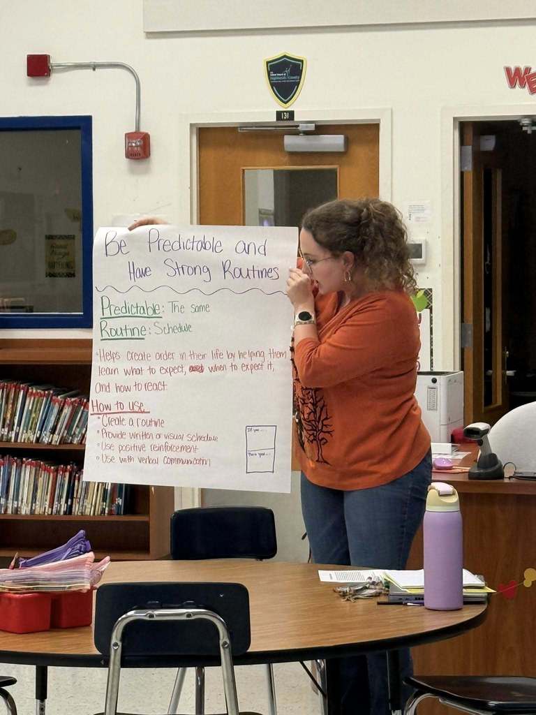 A woman with curly hair, wearing an orange shirt, stands next to a round table, holding and reading from a large poster board. The poster advises to "Be Predictable and Have Strong Routines." Bookshelves are visible on the left side of the room.