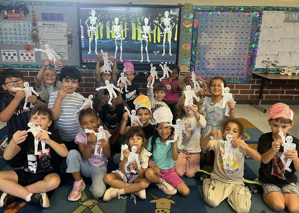 A diverse group of approximately fifteen elementary school children are sitting on a rug in a classroom, smiling and holding up paper skeletons they have made. A large screen behind them displays dancing skeleton figures, and classroom charts are on the walls.