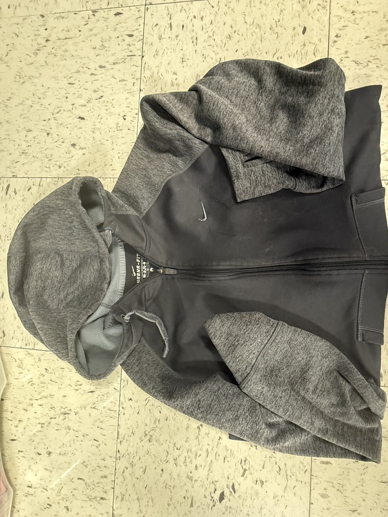 A black and gray Nike hoodie with a zipper is folded and lying on a tiled floor.