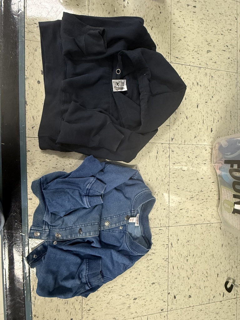Two folded items of clothing lay on a tiled floor: a dark navy or black hooded sweatshirt and a blue denim jacket with snap buttons.