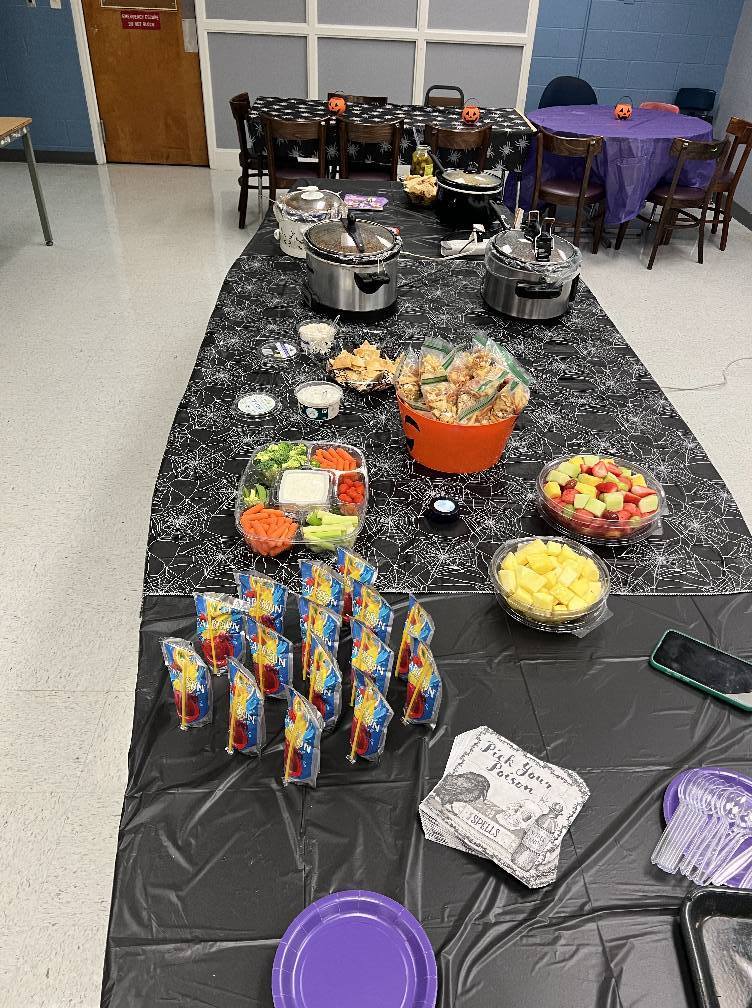 A long table set for a Halloween-themed party. It is covered with a black and white spiderweb cloth and features several slow cookers, a large vegetable tray with dip, bowls of fruit, bags of snacks, and purple plates and utensils. The tables in the background are also decorated for the event.