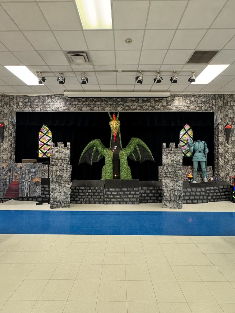 A stage is elaborately decorated for a medieval or fantasy theme, featuring a large green dragon centerpiece against a black curtain. The decor mimics a stone castle with turrets, stained-glass-like windows, a suit of armor statue, and a stone backdrop. The floor is white tile with a blue section.