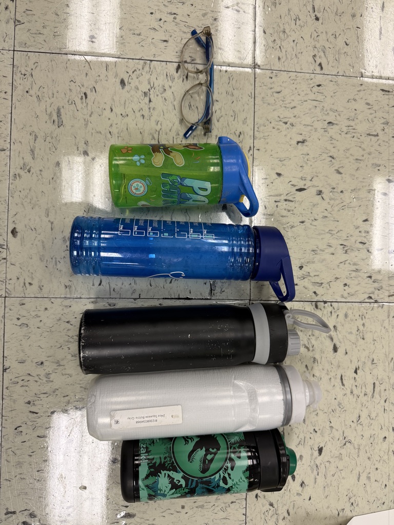 Five water bottles and a pair of blue-rimmed eyeglasses are lined up vertically on a tiled floor. The bottles include a small green one with Paw Patrol graphics, a tall blue one, a black insulated bottle, a plain white bottle, and a black one with a Jurassic Park design.