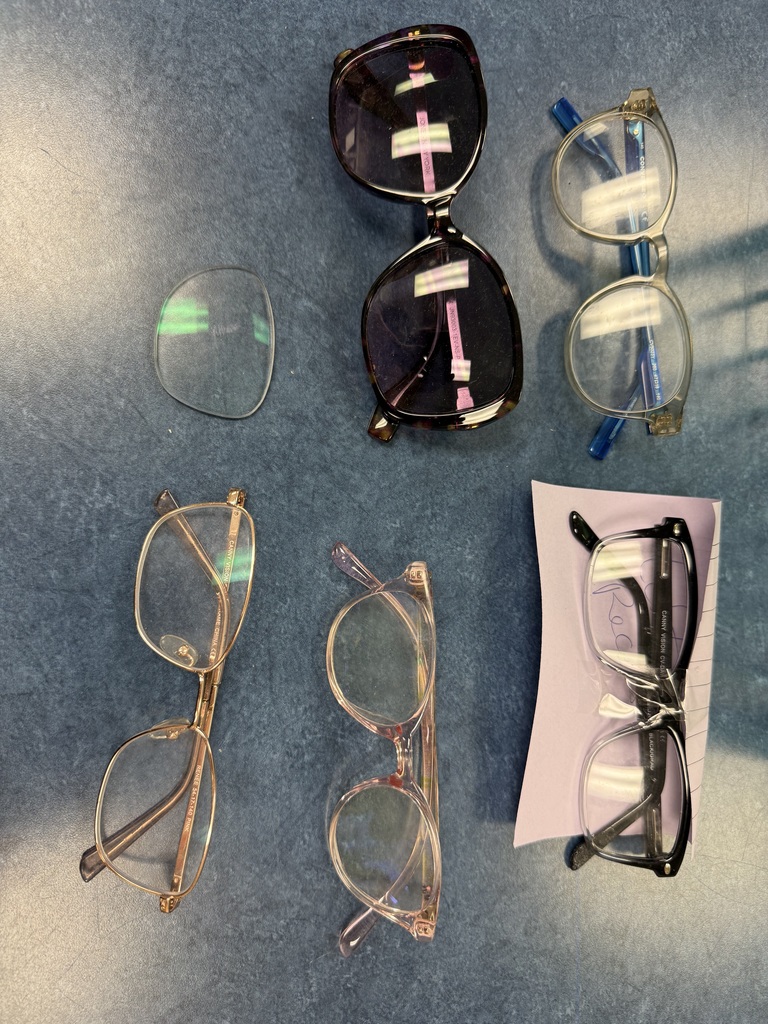 Six different pairs of eyeglasses and one detached lens are displayed on a blue surface. The glasses have various styles, including dark frames, clear/gold frames, and blue/silver frames.