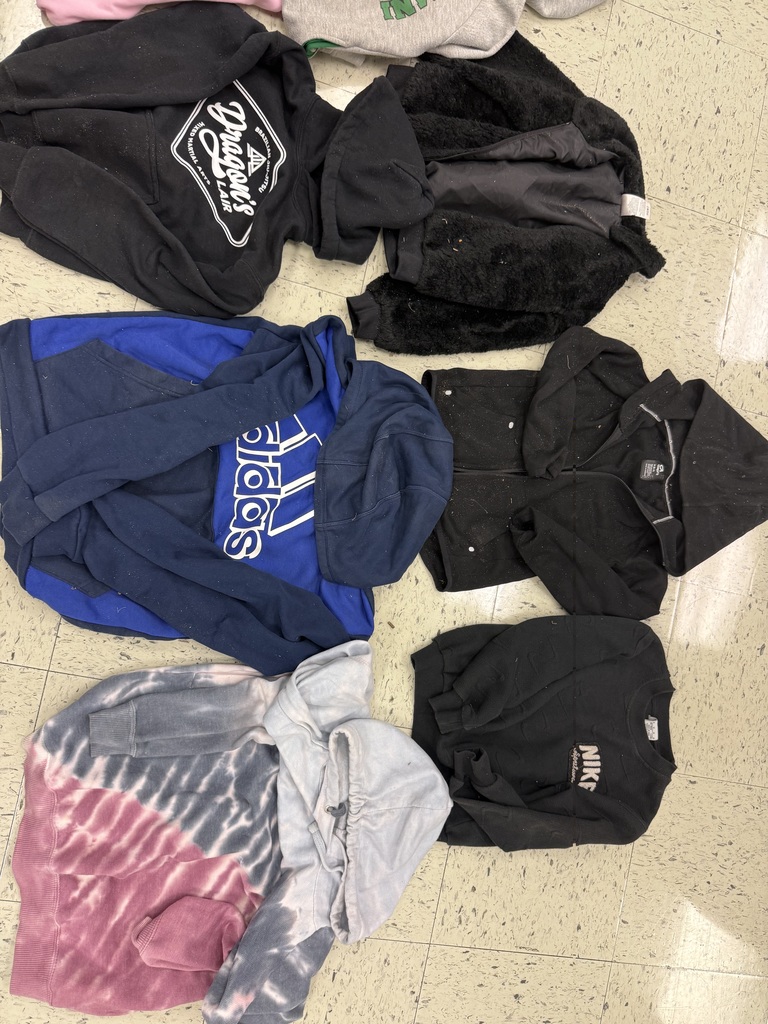 Seven dark-colored items of clothing are lying on a tiled floor. They include a black pullover, a black furry vest, a blue Adidas hoodie, a black zippered sweatshirt, a tie-dye hoodie, and two black long-sleeve shirts (one with "NIKE" on it).