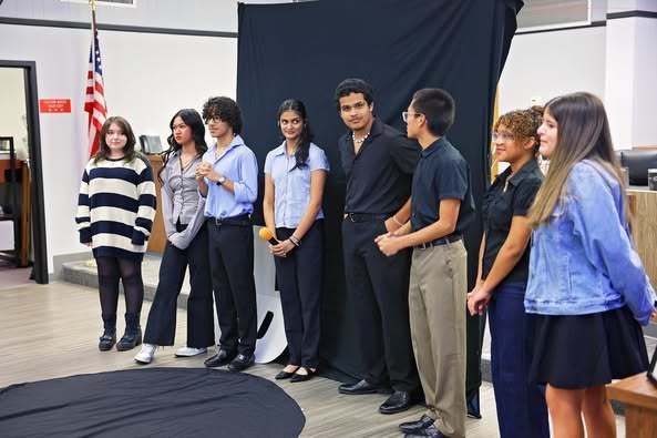 A diverse group of nine high school students is standing in front of a black backdrop with a logo on it, possibly for a presentation or performance. An American flag is visible on the left, and a black circular mat is on the floor in front of them.