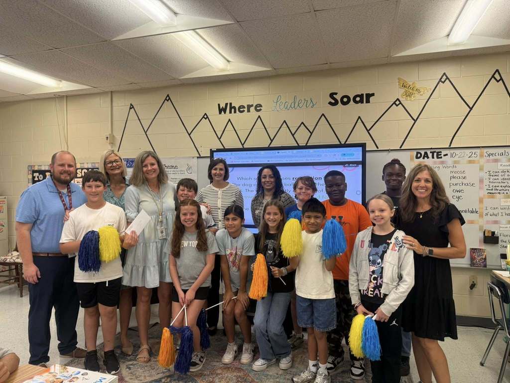A group of students holding blue and yellow pom-poms kneel or stand with several adults in front of an interactive whiteboard displaying educational content in a classroom.